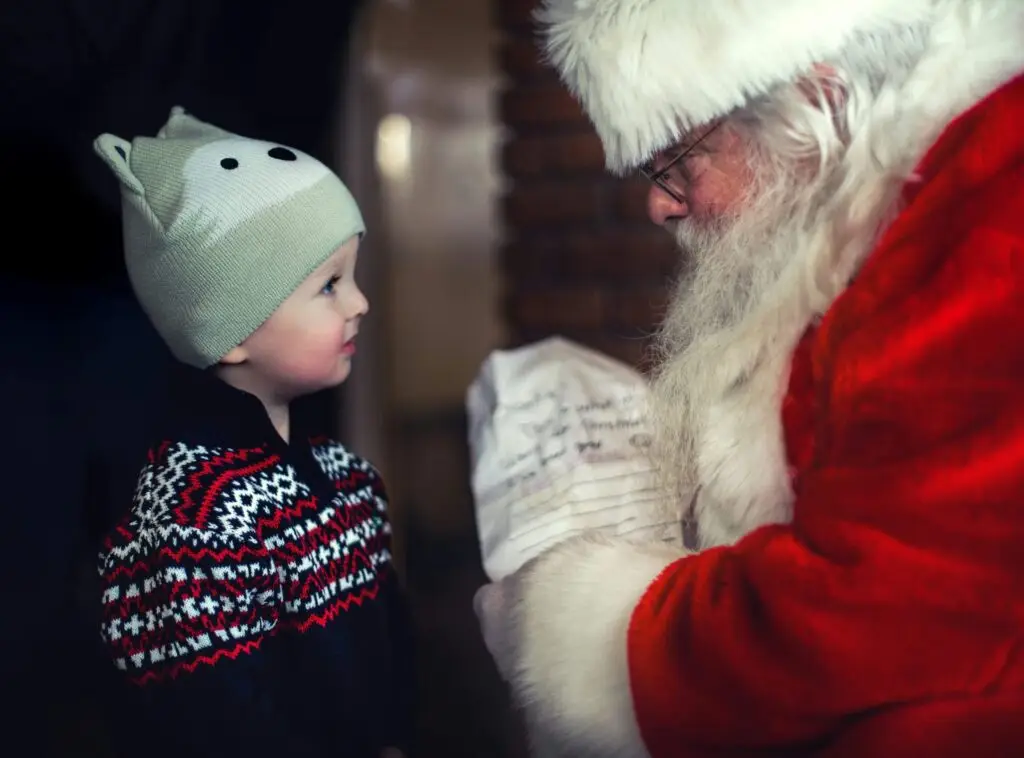 A young child wearing a winter hat with animal ears and a patterned sweater gazes at Jolly Old Saint Nicholas. Santa, in his red suit and hat, holds a letter and looks warmly at the child, creating a festive atmosphere.