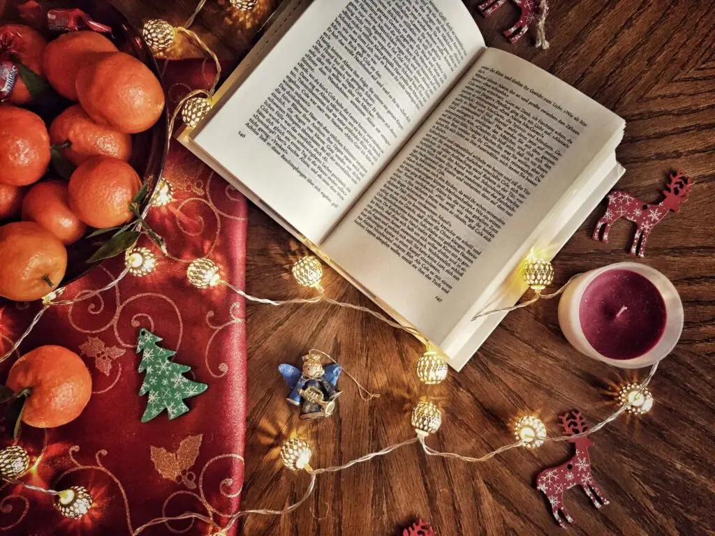 An open book on a wooden table reveals the history of Christmas, surrounded by festive decorations like string lights, red reindeer ornaments, a candle, and a bowl of tangerines resting on a red cloth adorned with green tree designs.