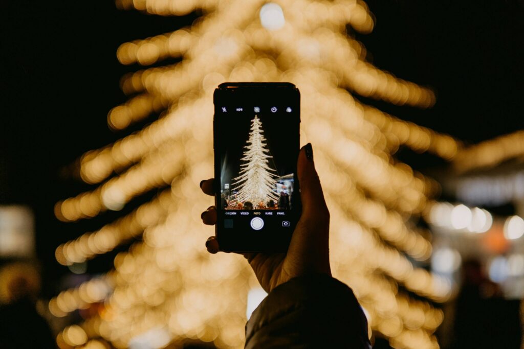 A person holds a smartphone, capturing a photo of a large, illuminated Christmas tree with glowing lights. The tree is blurred in the background while the phone's screen shows it in focus, providing inspiration for DIY Christmas wall art that brings festive cheer into your home.