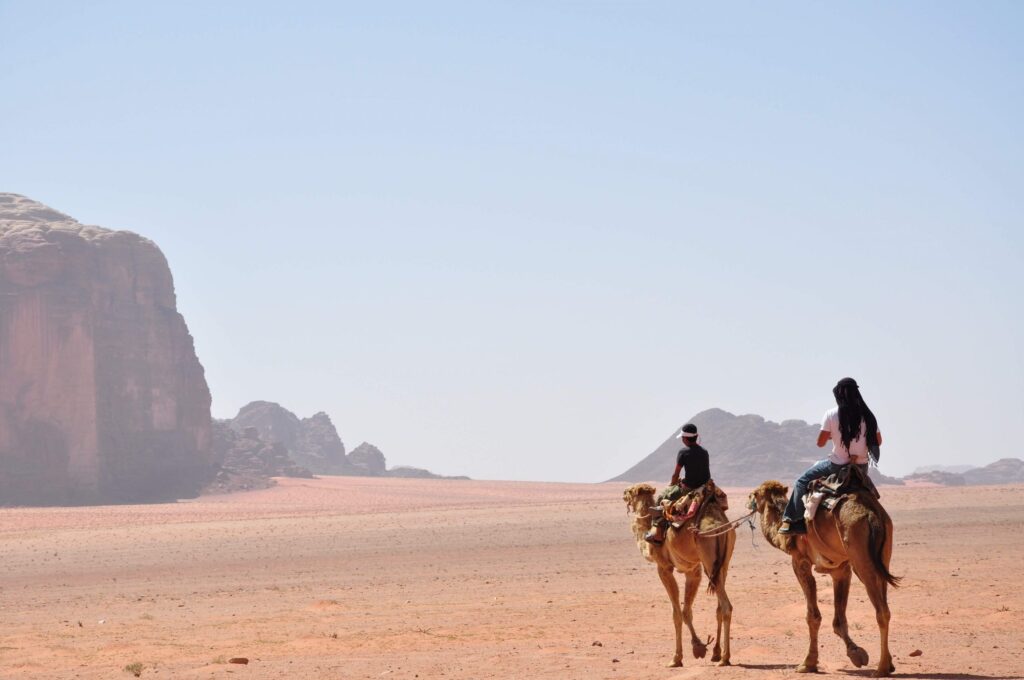 Two people ride camels across a vast desert landscape under a clear blue sky. Large rocky cliffs loom in the distance, creating a dramatic backdrop. The scene conveys a sense of adventure and exploration, perfect for those seeking packing tips for international travel.