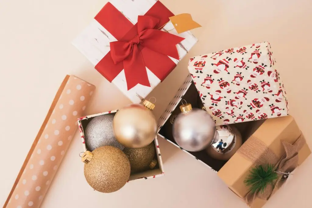 Gift boxes adorned with red ribbons and holiday-themed paper reveal DIY Christmas ornament kits in silver and gold. A roll of polka-dotted wrapping paper and a pine-decorated present are nearby, all set against a cream-colored background.