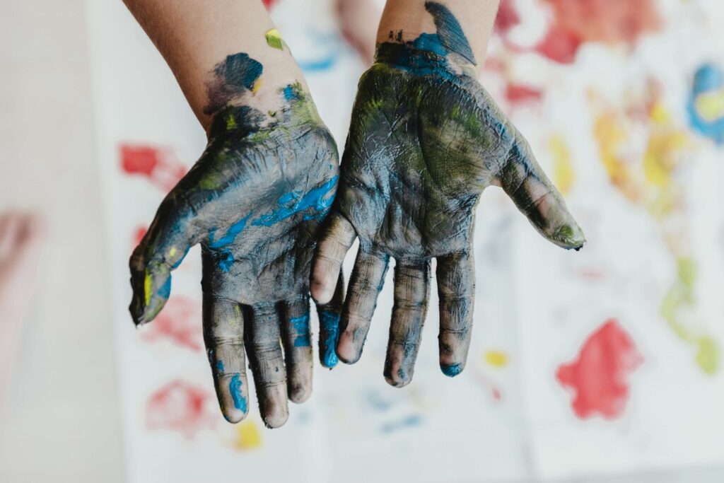 Two hands, covered in a festive mix of blues, greens, and blacks, form a heart shape against a backdrop where colorful paint splashes evoke the joyful chaos of Christmas spirit on a white canvas.