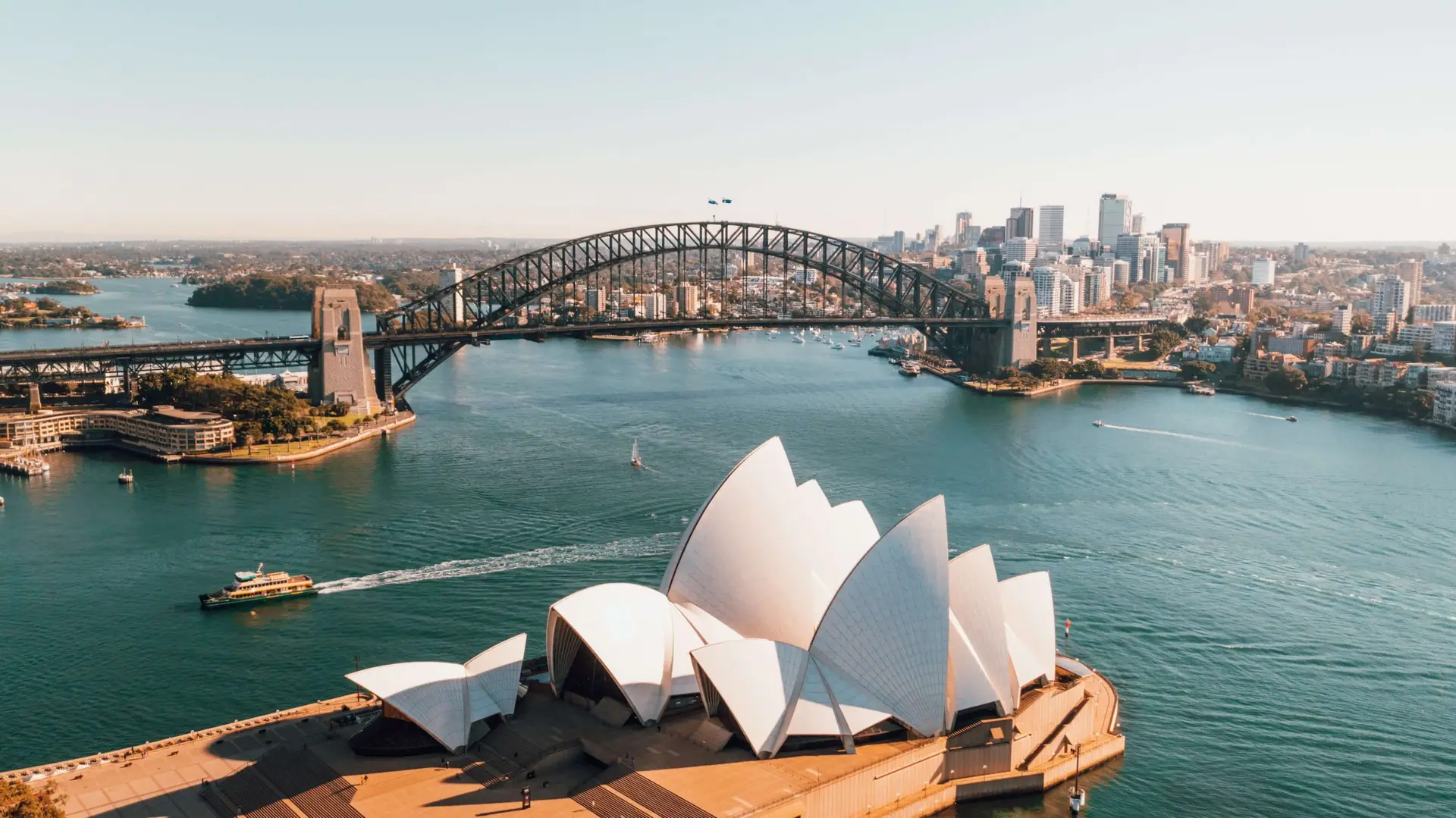 Aerial view of the Sydney Opera House with its distinct white sails in the foreground. The Sydney Harbour Bridge arcs majestically behind, spanning across the harbor where Christmas in Australia is celebrated under the summer sun. The cityscape of Sydney shimmers beneath a clear blue sky.