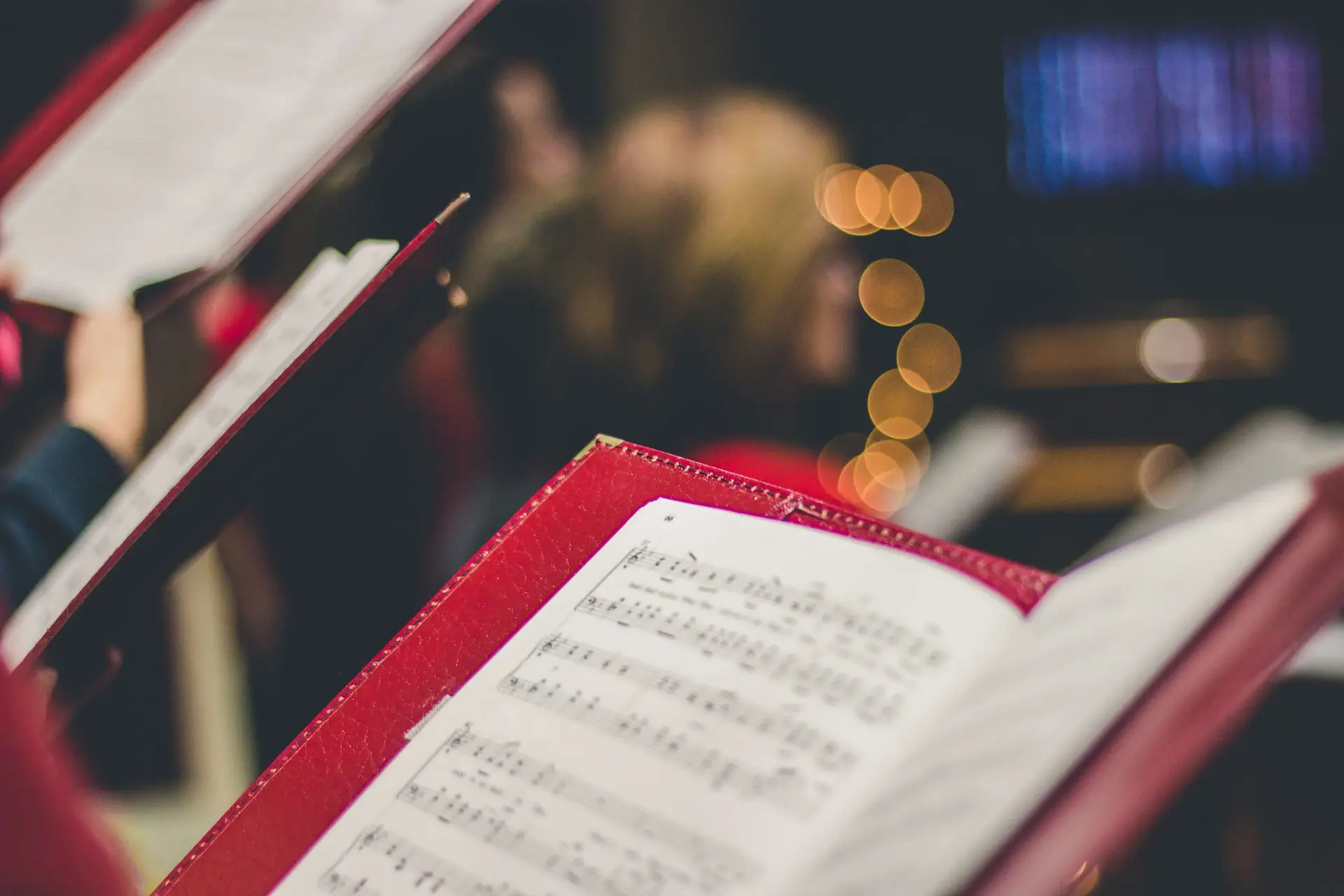 Close-up of open music books with sheet music, held by choir members during a performance. The background is softly blurred, showcasing bokeh lights and a hint of audience or choir members. This captures the rich tradition and history of Christmas carols celebrated through song.