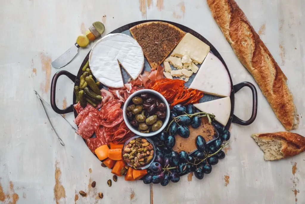 A festive Christmas charcuterie board adorned with cured meats, cheeses, olives, grapes, pistachios, and vegetables. A sliced baguette sits invitingly on the side with a small knife nearby. The board rests on a rustic white wooden surface.