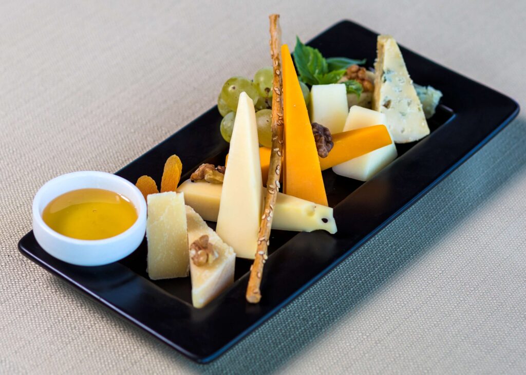 A rectangular black plate offers a delightful Christmas snack mix featuring assorted cheese slices, walnuts, grapes, dried apricots, and a breadstick. A small white bowl of olive oil sits on the side, all set against a light textured fabric background.