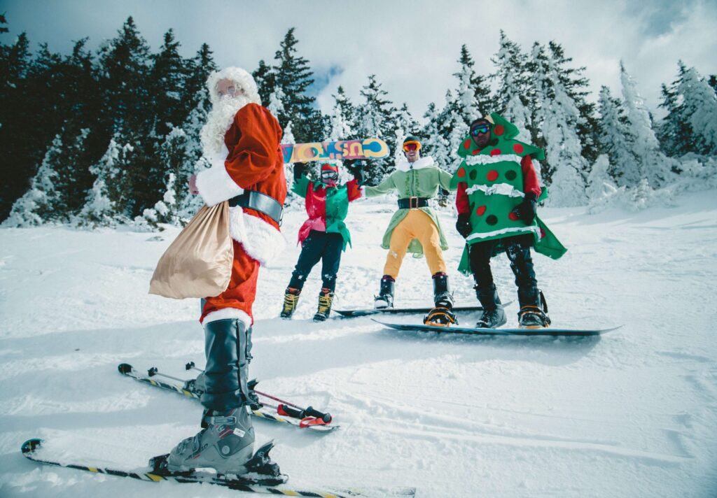 A group of four in festive Christmas party outfits is snowboarding and skiing on a snowy hill. One, dressed as Santa, skis while the others, dressed as a Christmas tree, elf, and gift, stand with snowboards. Snow-covered trees form the backdrop.