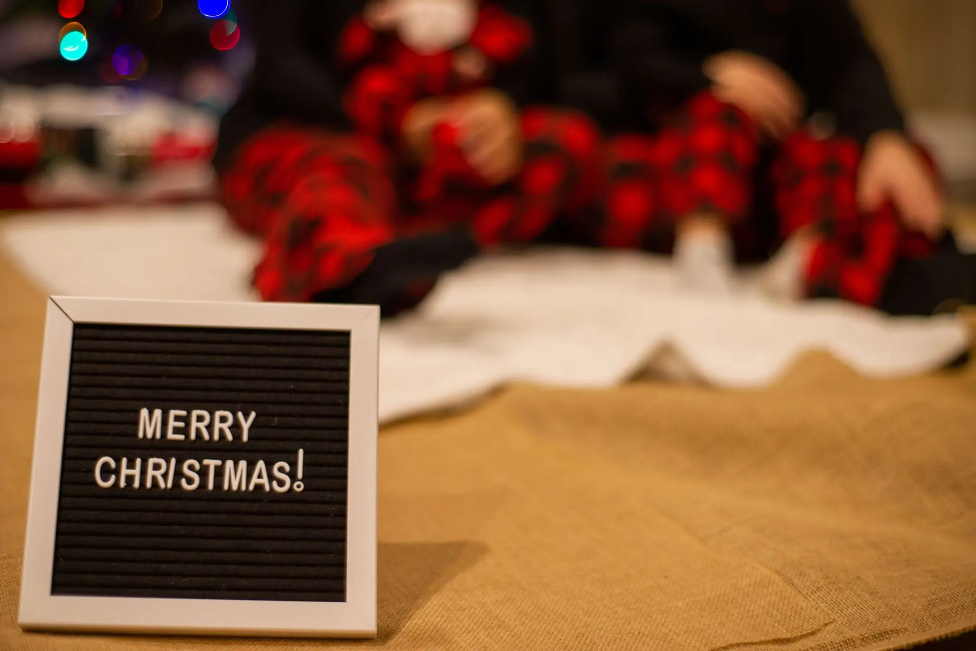 A felt letter board with Merry Christmas! takes center stage, while in the blurred background, two people dressed for a Christmas pajama party in red and black plaid snuggle together on a cozy blanket.