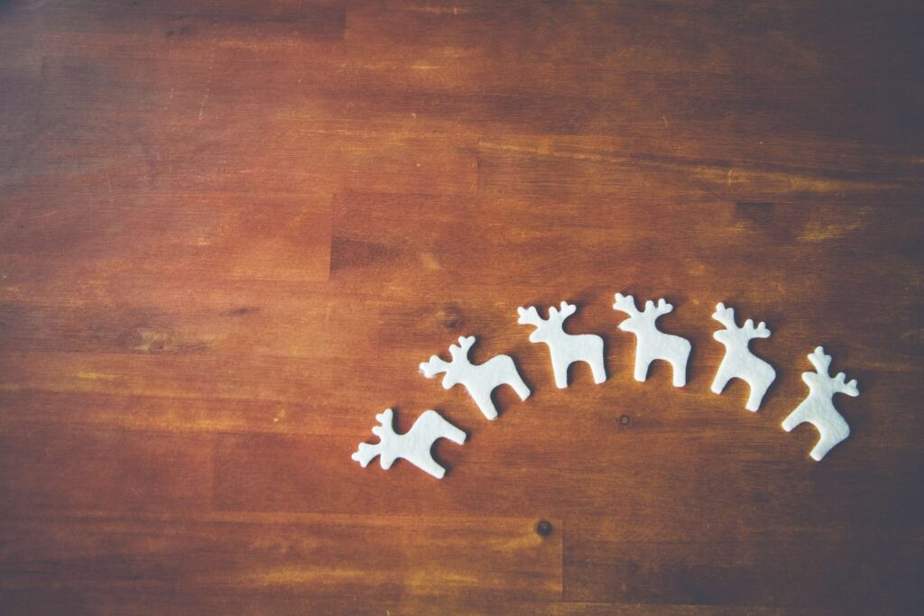 Five small, white reindeer figurines are arranged in a curved line on a wooden surface, surrounded by DIY felt Christmas ornaments. The background is a warm, brown texture, creating a cozy and festive atmosphere.