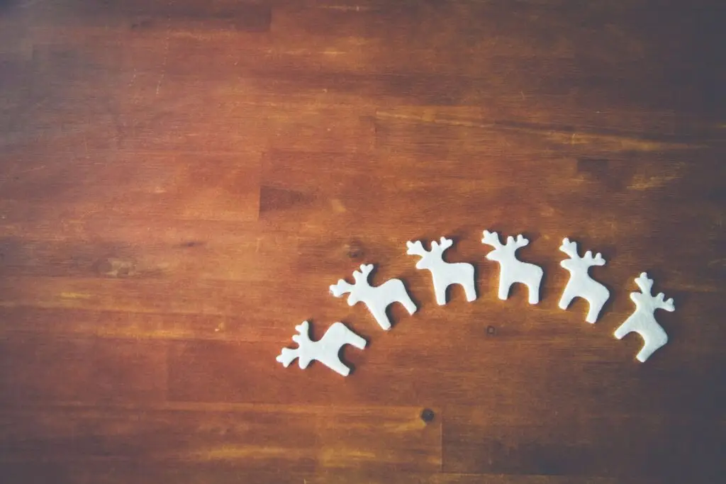 Five small, white reindeer figurines are arranged in a curved line on a wooden surface, surrounded by DIY felt Christmas ornaments. The background is a warm, brown texture, creating a cozy and festive atmosphere.