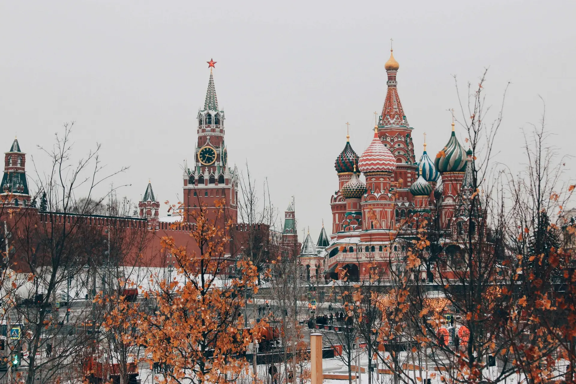 A view of the Moscow Kremlin and Saint Basil's Cathedral in winter showcases the red brick walls and colorful onion domes against a cloudy sky. Bare trees and snow patches foreground the scene where fun facts about Christmas in Russia often include festive traditions echoing from these iconic landmarks.