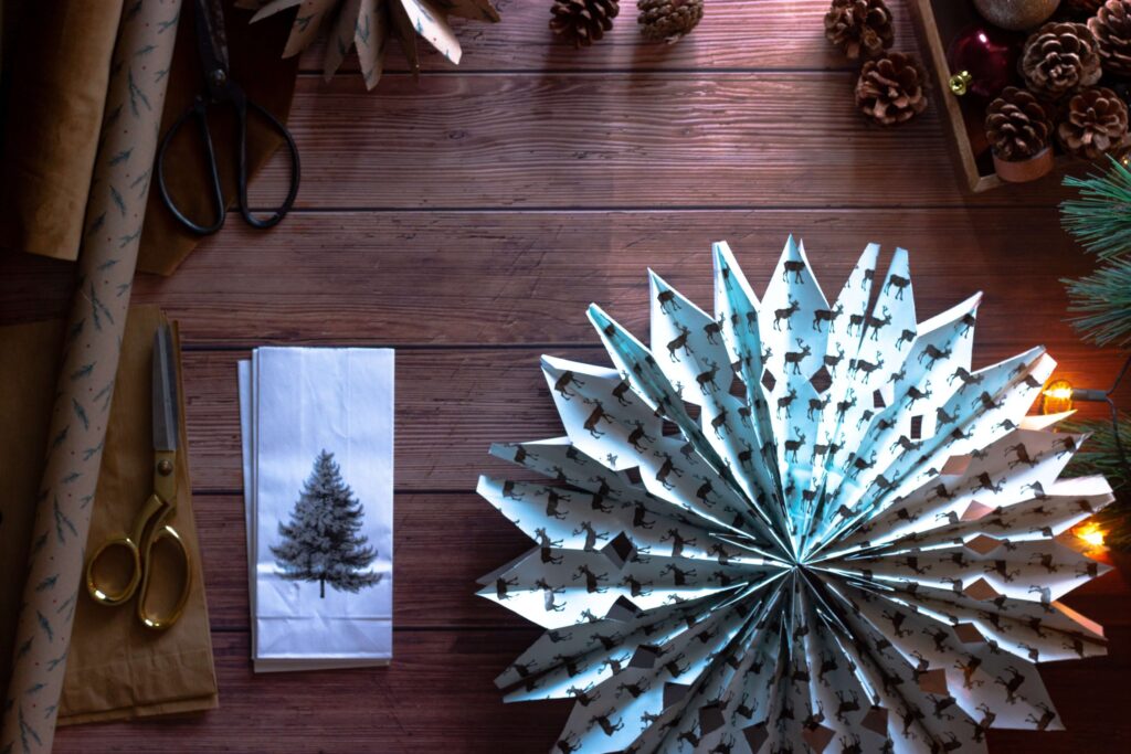 A festive wooden table adorned with paper snowflakes, pinecones, and string lights sets the perfect backdrop for crafting DIY Christmas ornaments for kids. Scissors and wrapping paper await creativity, while a folded napkin with a tree design and a Christmas tree branch complete the holiday scene.