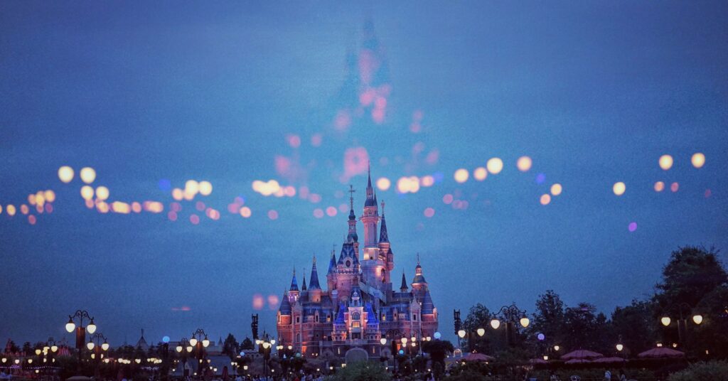 An illuminated castle stands at twilight, enveloped in a magical atmosphere akin to stepping into a Disney Christmas dream. Bokeh lights float above, casting a whimsical glow. The sky is deep blue, with trees and lamp posts completing this enchanting scene.