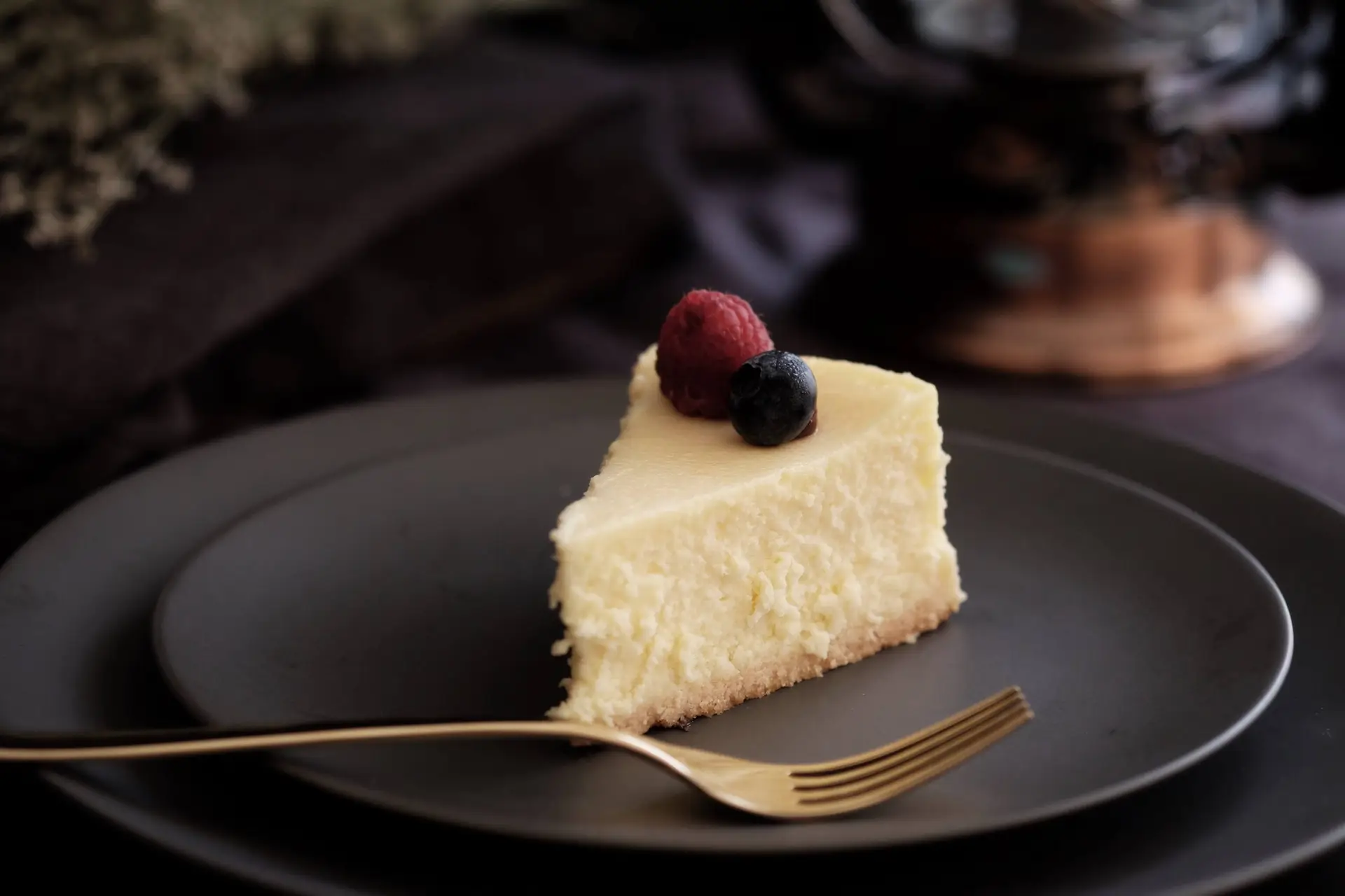 A slice of festive cheesecake graces a dark plate, topped with a raspberry and blueberry. A gold fork rests nearby, embodying the elegance of Christmas cheesecake recipes. The softly lit background creates a cozy and elegant atmosphere perfect for the holiday season.