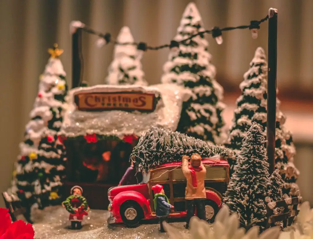 A festive Christmas scene with a red truck carrying a tree, surrounded by snow-dusted pine trees. Small figurines, including a person adjusting the tree and another holding a wreath, inspire cozy winter holiday vibes and spark creative Christmas party ideas for kids.