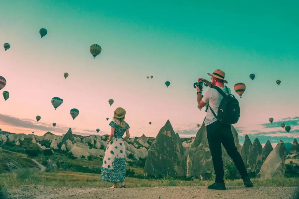 A man and woman stand on a rocky landscape at sunset, where numerous hot air balloons float in the sky. The man snaps photos, while the woman looks on, both dressed casually with hats. The greenish tint of the sky adds a dreamlike quality, reminiscent of travel toys for kids that inspire adventure.