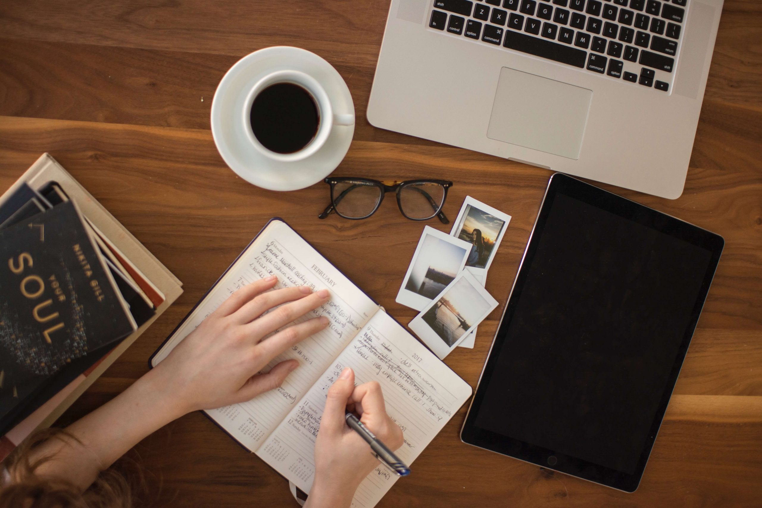 person writing in calendar journal with computer and ipad next to her