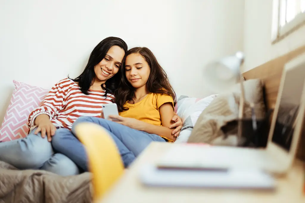 mom and daughter sitting on a bed looking at a smartphone together