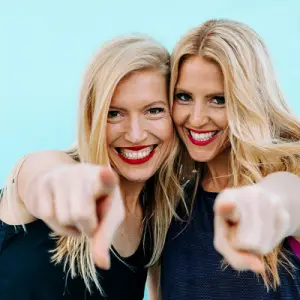 Two women with long blonde hair are smiling and pointing towards the camera. They are wearing sleeveless tops and standing against a bright blue background. Both have red lipstick and appear cheerful and friendly.