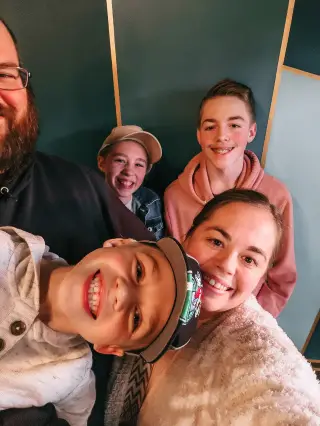 A smiling family of five taking a selfie against a green and gold background. A man with a beard is partly visible, while two boys and a girl wearing hats are looking into the camera. The woman in front is holding the camera.