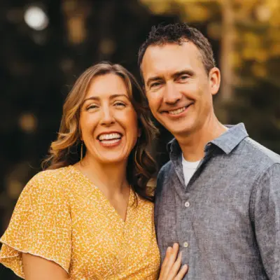 A smiling couple stands close together outdoors. The woman is wearing a yellow patterned dress, and the man is in a blue button-up shirt. The background is softly out of focus, featuring greenery and warm lighting.