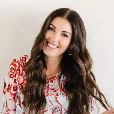 A woman with long, wavy brown hair smiles at the camera. She is wearing a colorful, patterned blouse with red and blue designs. The background is a plain, light-colored wall.