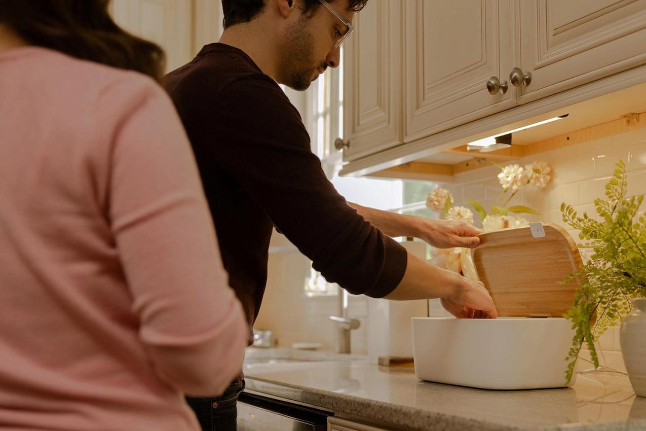 A man wearing glasses places items into a wooden container on a kitchen counter. A woman in a pink sweater stands nearby. The kitchen is brightly lit, with white cabinets and a vase of flowers.