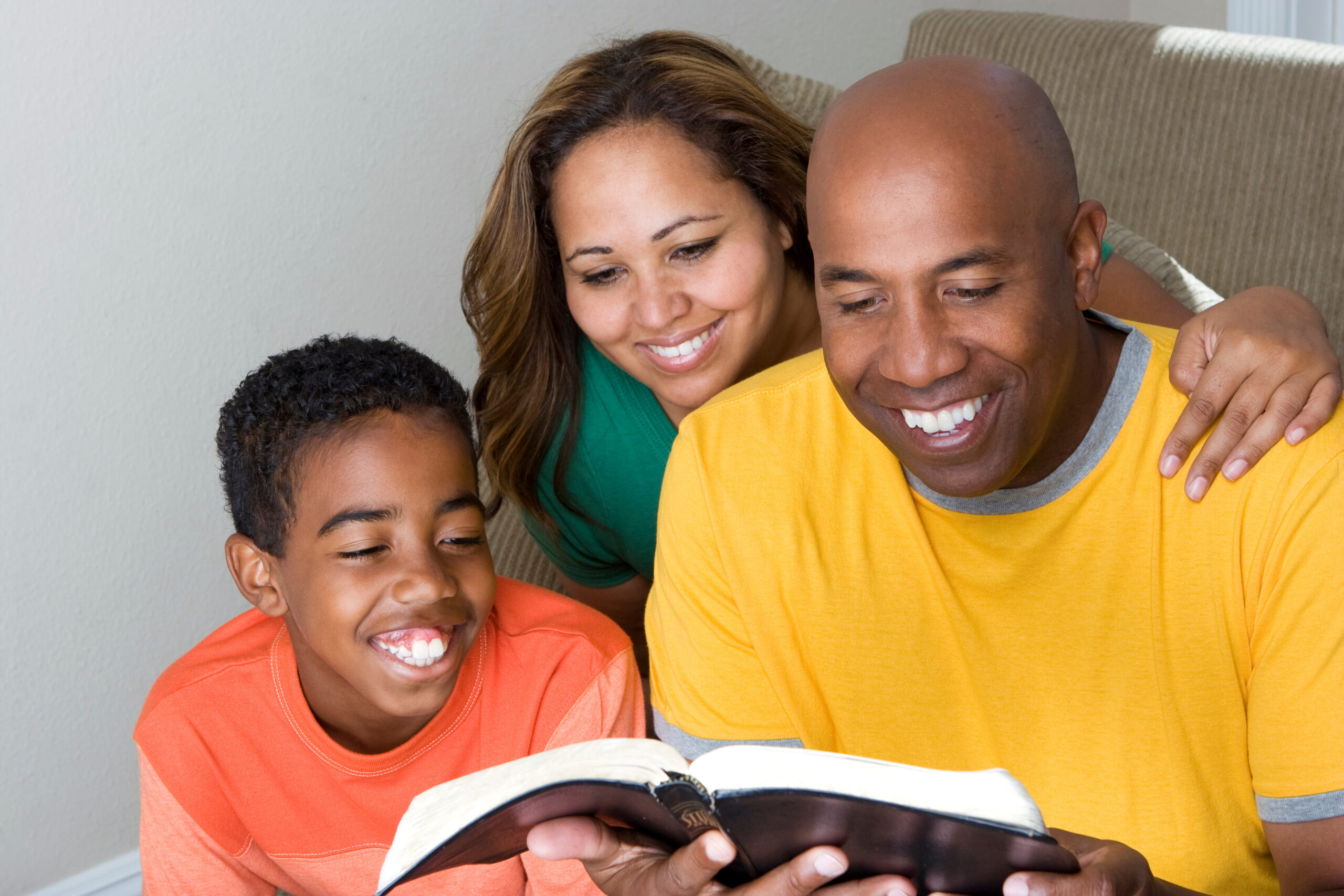 A family of three sits together on a couch, smiling while reading a book. The father, in a yellow shirt, holds the book as the mother, in a green shirt, and the son, in an orange shirt, look on. They appear happy and engaged.