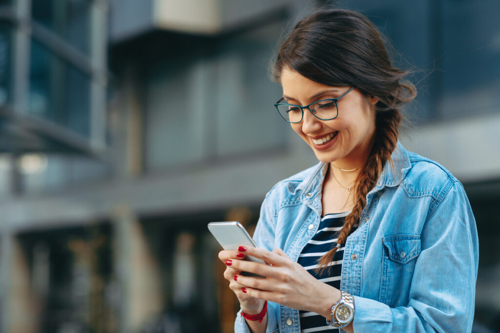 A woman wearing glasses and a denim jacket is smiling while looking at her smartphone. She stands outdoors in front of a modern building with large windows.