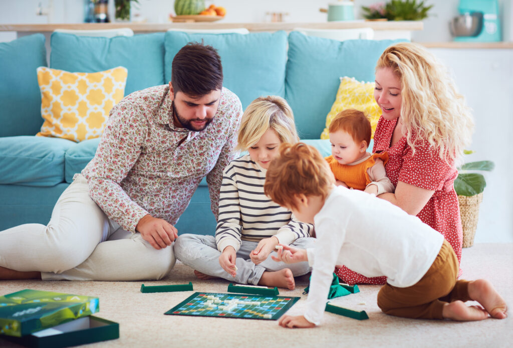 A family of five, including a baby and young children, is sitting on the floor in a cozy living room. They are playing a board game together. A blue sofa and colorful pillows are in the background. Everyone looks engaged and happy.