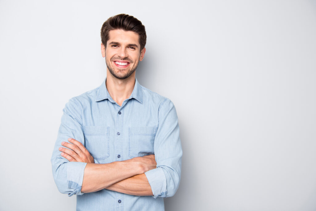 A man in a light blue button-up shirt stands against a light gray background, smiling with arms crossed.