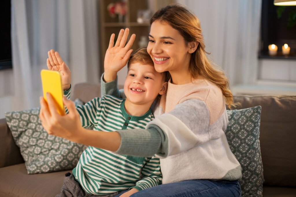 A woman and a young boy sit on a couch, smiling and waving at a yellow smartphone theyre holding up. They appear to be engaged in a video call. The room is warmly lit, with patterned cushions and candles in the background.