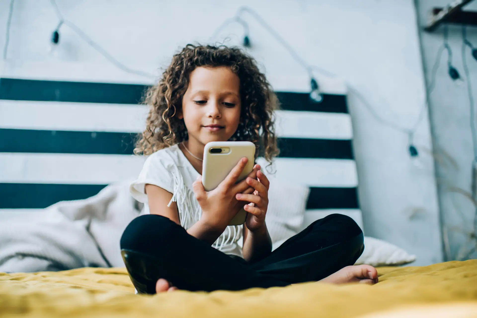 A young girl with curly hair is sitting cross-legged on a bed, holding a smartphone. She is focused on the screen. The room has a casual decor with black and white stripes on the headboard and string lights in the background.