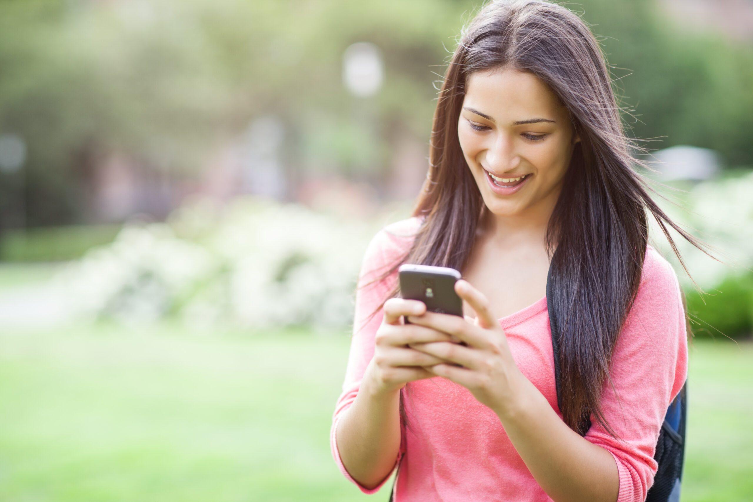 A young woman with long dark hair smiles while looking at her smartphone. She is wearing a pink top and standing outdoors in a park-like setting, with green grass and blurred trees in the background.