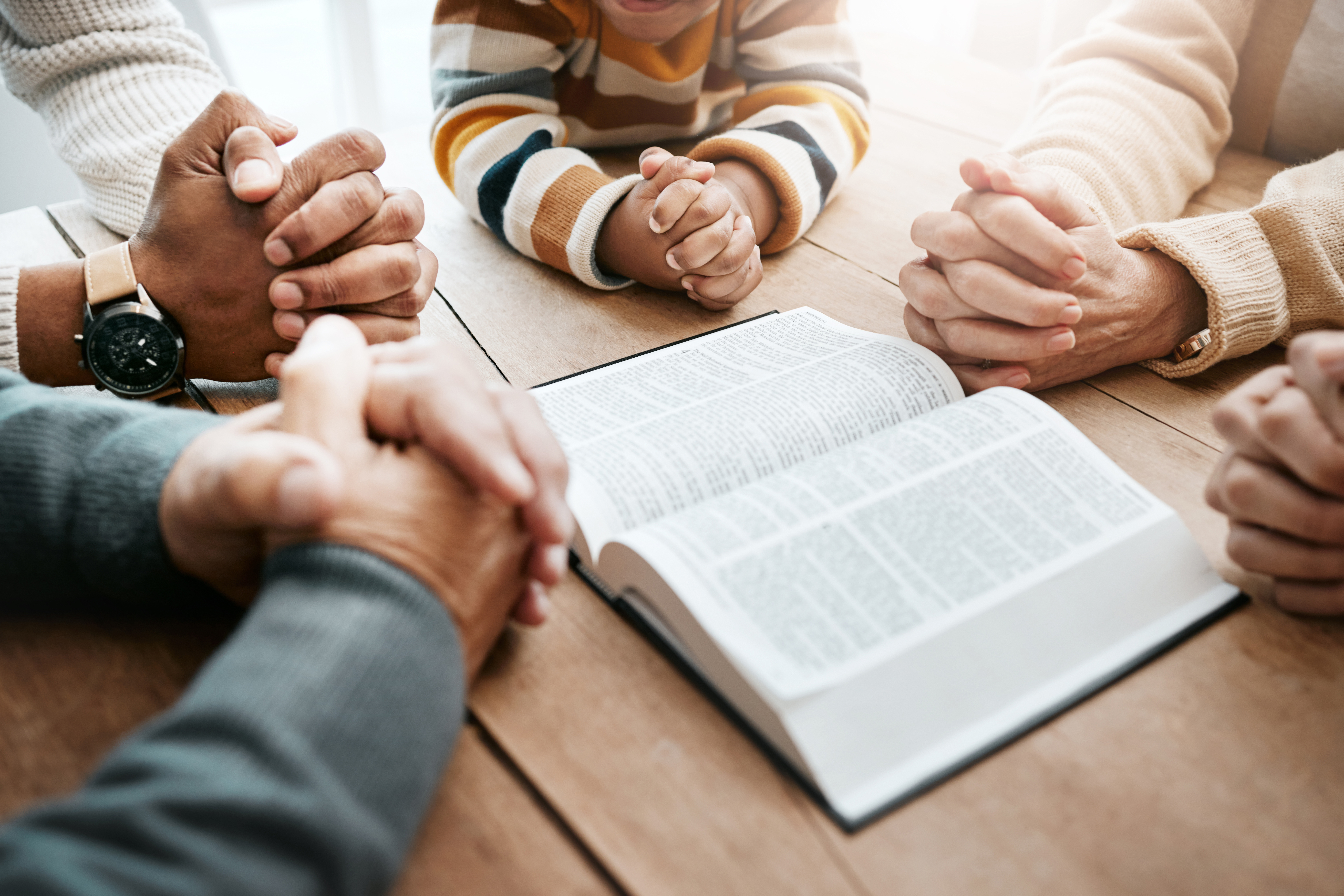 A group of people sit around a wooden table with their hands clasped in prayer. An open book, possibly a Bible, is in the center. They are wearing cozy sweaters and one person has a striped shirt. Sunlight gently illuminates the scene.