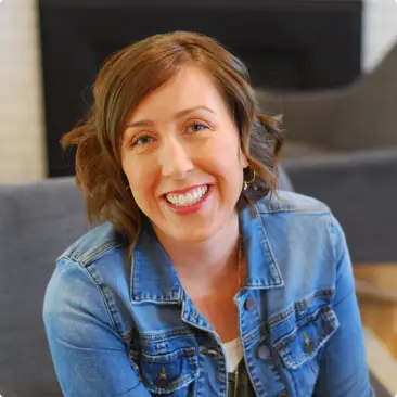 A smiling person with short brown hair sits in a cozy room wearing a denim jacket. A fireplace and gray chair are visible in the background.