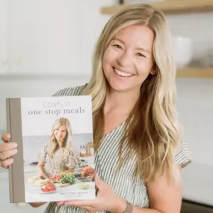 A woman with long blonde hair smiles while holding a cookbook titled Costco One Stop Meals. She is wearing a striped shirt and stands in a kitchen.