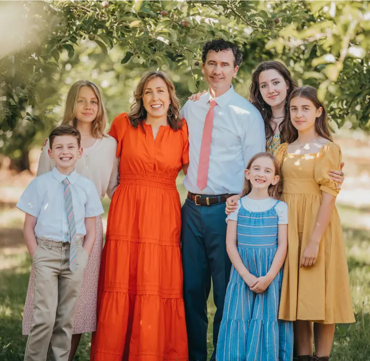 A family of seven poses outdoors under leafy trees. The mother and father stand in the center, surrounded by five children, three girls and two boys. They are all dressed in bright, summery clothes and smiling at the camera.