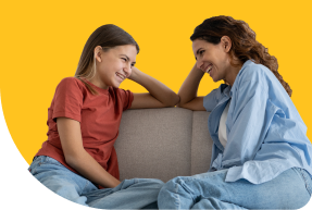 Two people, one wearing a red shirt and the other a blue shirt, are sitting on a couch facing each other, smiling and talking. The background is a solid yellow color.