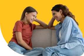 Two people, one wearing a red shirt and the other a blue shirt, are sitting on a couch facing each other, smiling and talking. The background is a solid yellow color.