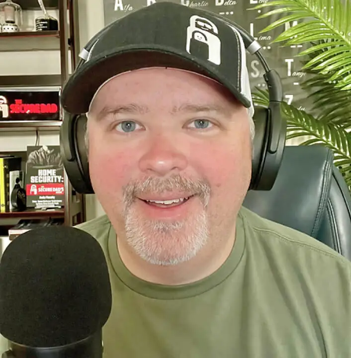 A person wearing a black cap and headphones, speaking into a microphone. They are sitting in a room with a visible plant and bookcase in the background.