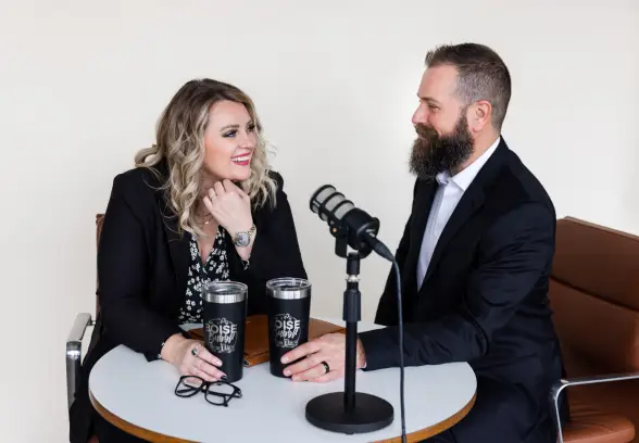 A woman and a man are seated at a round table with a microphone. They are engaged in conversation, smiling, each holding a black tumbler. The woman has glasses and wears a black jacket, while the man has a beard and is in a suit.