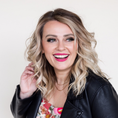 A woman with wavy blonde hair smiles brightly. She is wearing a black leather jacket over a floral top. The background is a plain, light color.