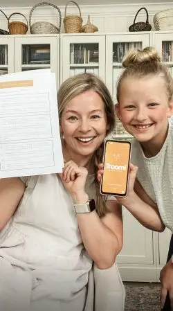 A woman and a child smile at the camera. The woman holds a set of documents, while the child holds a smartphone displaying a screen with the text Troomi. They are in a room with white cabinets and a bookshelf in the background.