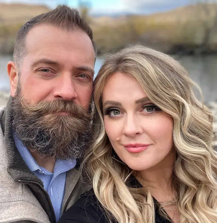 A couple taking a selfie outdoors near a river. The man has a beard and short hair, while the woman has long, wavy blond hair. They are both wearing jackets, and the background features water and a blurred landscape of hills and trees.