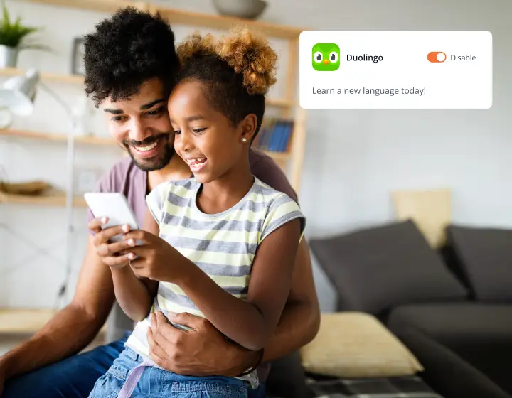 A father and daughter sit on a couch, happily looking at a smartphone together. A notification from Duolingo on the screen says, Learn a new language today! The room is bright and cozy.