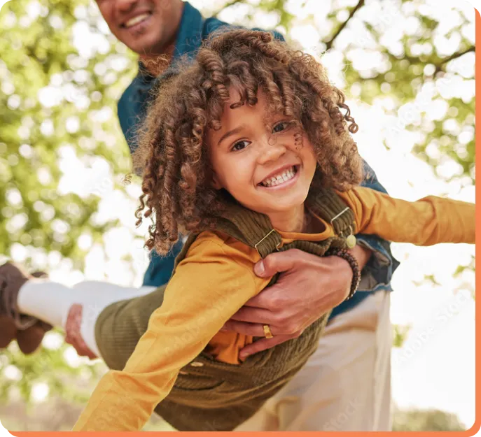 A child with curly hair smiles brightly while being spun around playfully by an adult outside. The child is wearing yellow and green, and the background shows trees and sunlight, creating a warm, joyful atmosphere.