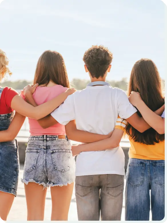 Four people stand close together with their backs to the camera, arms wrapped around each others shoulders. They are outdoors, wearing casual summer clothes, and appear to be looking at a beach or seaside view under a clear blue sky.