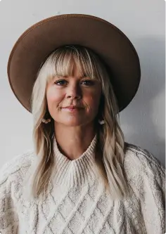 A woman with long blonde hair and bangs is wearing a wide-brimmed brown hat and a cozy white cable-knit sweater, standing against a plain white background. She is looking directly at the camera with a neutral expression.