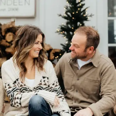 A couple sits closely in front of a decorated Christmas tree, smiling at each other. The woman wears a patterned sweater, and the man is in a brown shirt. A stack of firewood is visible in the background.